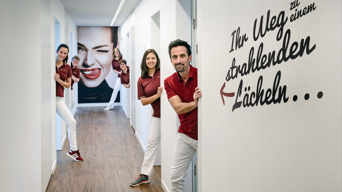Team der Praxis Dr. Wingendorf im Flur - an der Wand der Spruch "Ihr Weg zu einem strahlenden Lächeln"