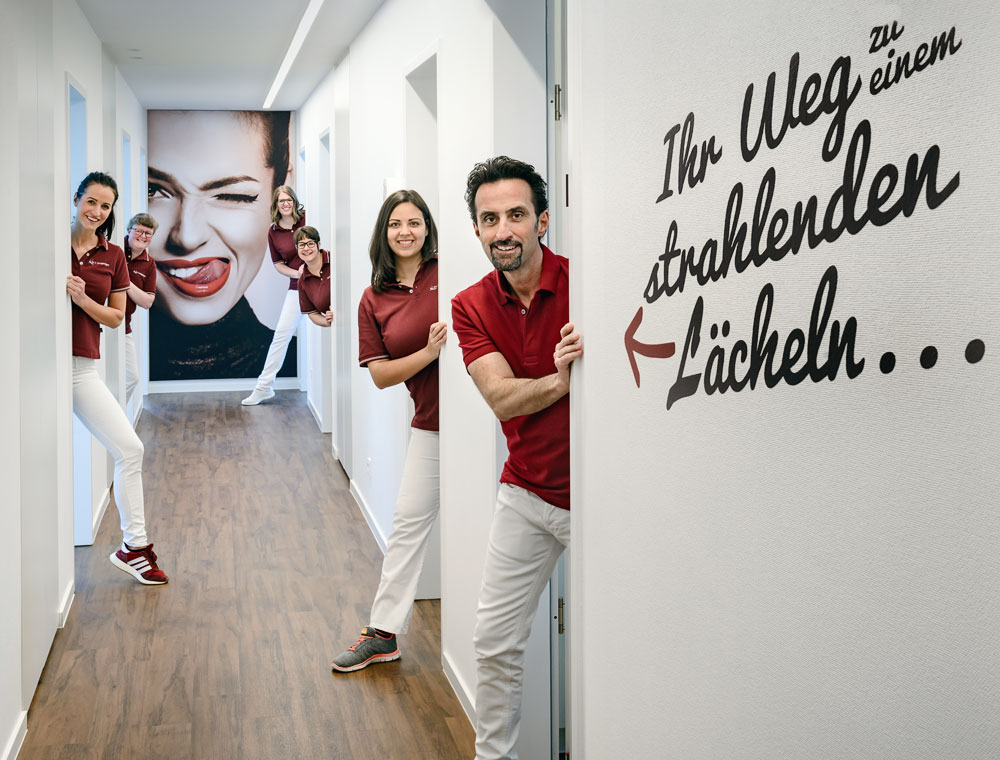 Team der Praxis Dr. Wingendorf im Flur - an der Wand der Spruch "Ihr Weg zu einem strahlenden Lächeln"